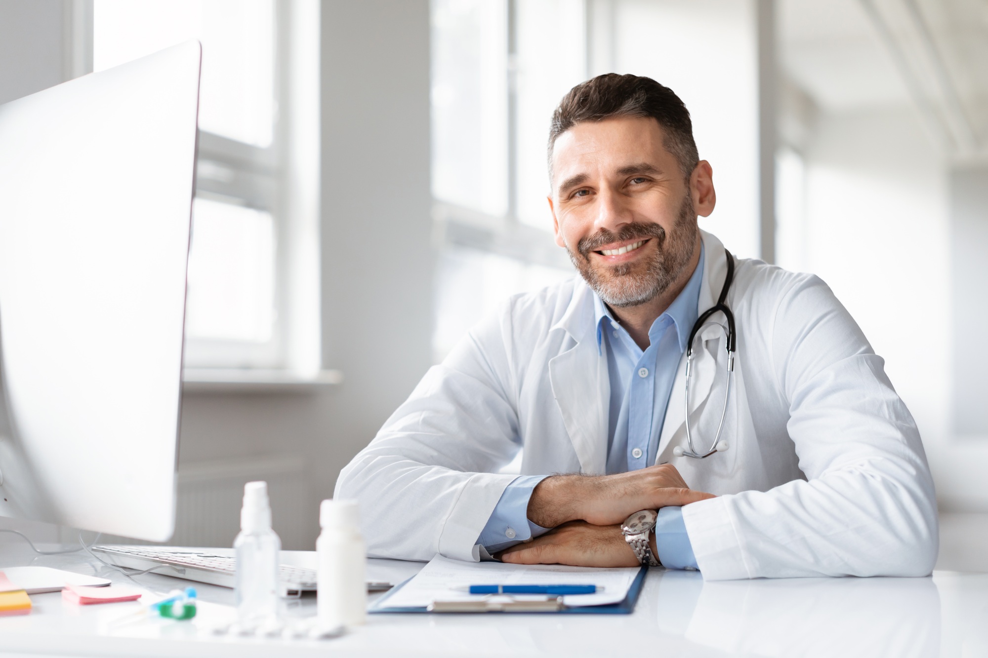 Portrait of happy male doctor in white medical uniform, sitting at desk in hospital and smiling at