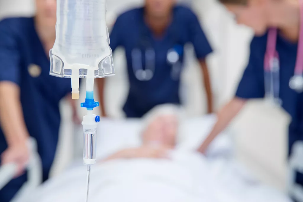 Perfusion intraveineuse au premier plan tandis qu’une équipe médicale accompagne un patient hospitalisé, illustrant les enjeux de la fin de vie pour les soignants.