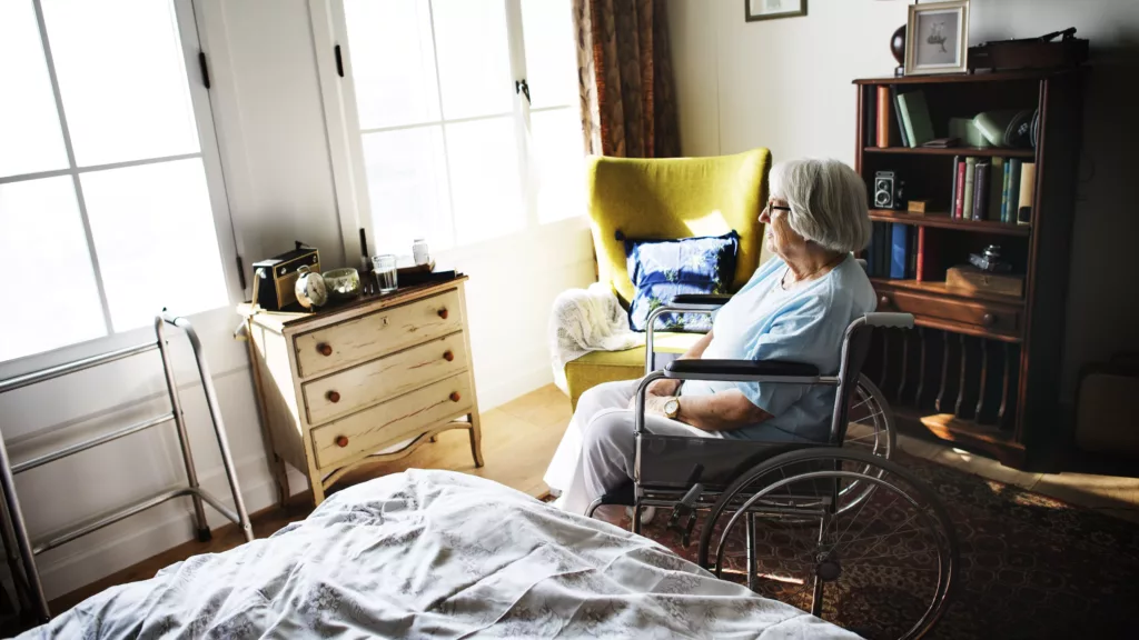 Femme senior en fauteuil roulant seule chez elle regardant par la fenêtre, situation de perte d’autonomie